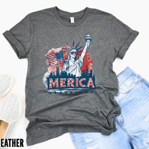 May include: A deep heather gray t-shirt featuring a graphic of the Statue of Liberty wearing sunglasses, an American flag, and the word "MERICA" in red, white, and blue. The shirt is paired with denim shorts and white slip-on shoes.