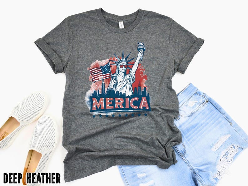 May include: A deep heather gray t-shirt featuring a graphic of the Statue of Liberty wearing sunglasses, an American flag, and the word "MERICA" in red, white, and blue. The shirt is paired with denim shorts and white slip-on shoes.