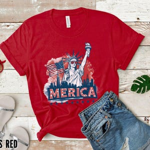 May include: Red t-shirt with a graphic of the Statue of Liberty, an American flag, and the word "MERICA". The shirt is paired with denim shorts, red sunglasses, and silver sandals. The words "CANVAS RED" are at the bottom.
