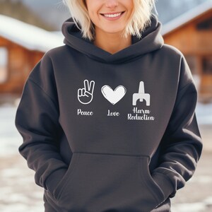 May include: Dark gray hoodie with the words "Peace Love Harm Reduction" in white. The design includes a peace sign, a heart, and a stylized symbol. The hoodie has a drawstring hood and a front pocket.