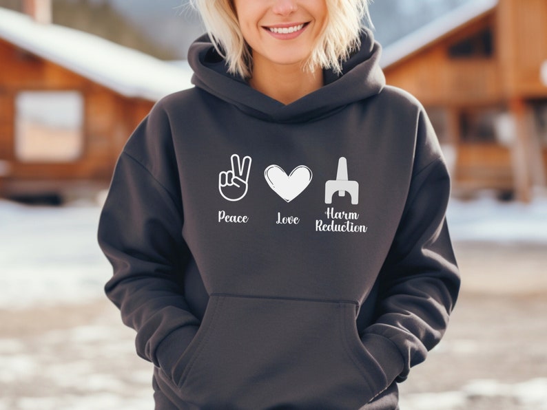 May include: Dark gray hoodie with the words "Peace Love Harm Reduction" in white. The design includes a peace sign, a heart, and a stylized symbol. The hoodie has a drawstring hood and a front pocket.
