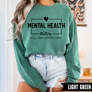 Peut inclure: Sweat-shirt vert clair avec le texte "MENTAL HEALTH Matters ALL DAY EVERY DAY" en noir. Le motif comprend un c&oelig;ur noir au-dessus du mot "MENTAL HEALTH". Le sweat-shirt est associ&eacute; &agrave; un short en jean d&eacute;lav&eacute;.