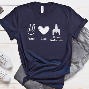 May include: Navy blue t-shirt with white graphic symbols for peace, love, and harm reduction. The text "Peace Love Harm Reduction" is printed below the symbols. The shirt is paired with gray jeans, white sneakers, and sunglasses.
