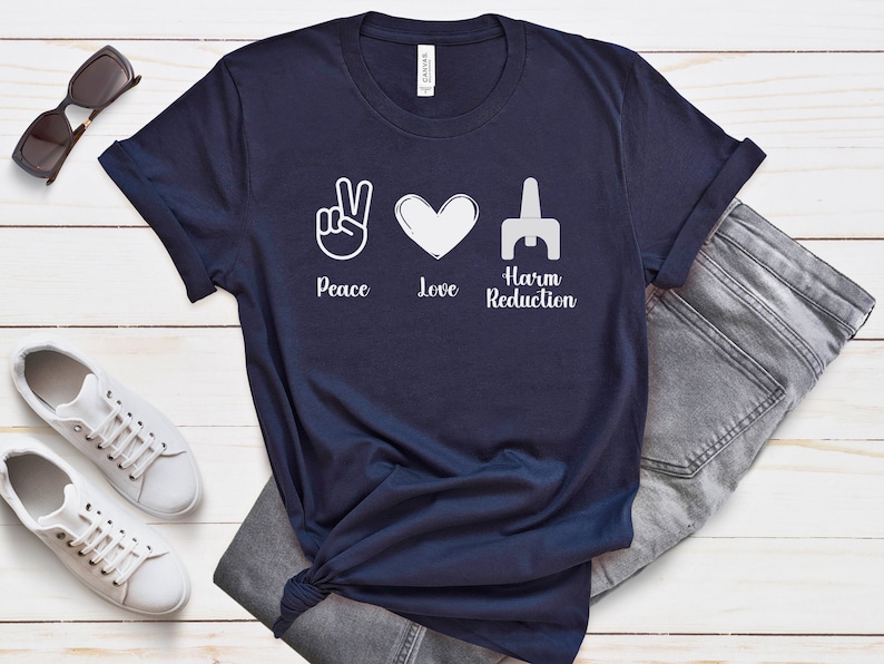 May include: Navy blue t-shirt with white graphic symbols for peace, love, and harm reduction. The text "Peace Love Harm Reduction" is printed below the symbols. The shirt is paired with gray jeans, white sneakers, and sunglasses.