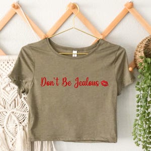 Puede incluir: Una camiseta verde recortada con el texto "Don't Be Jealous" en letras rojas y una impresi&oacute;n de labios rojos.
