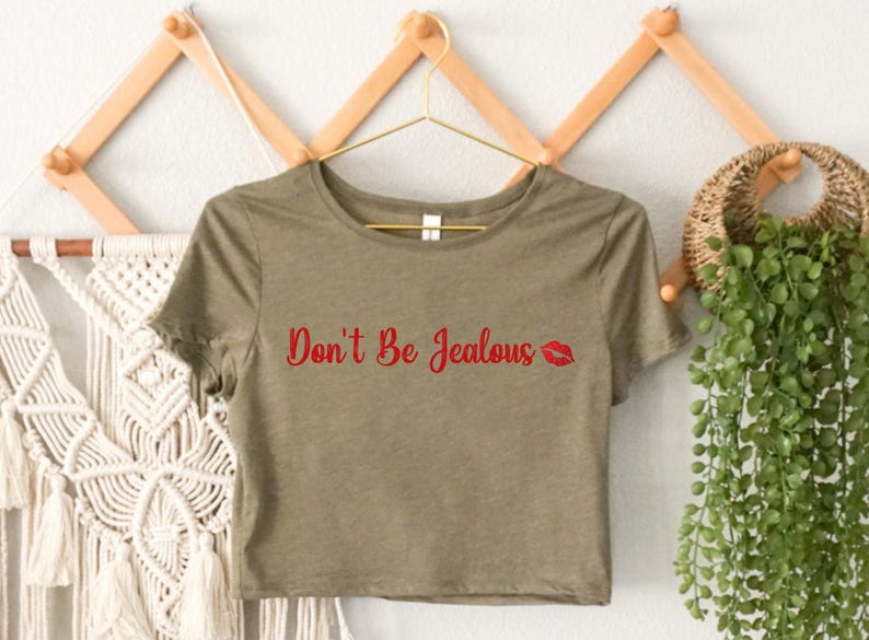 Puede incluir: Una camiseta verde recortada con el texto "Don't Be Jealous" en letras rojas y una impresi&oacute;n de labios rojos.