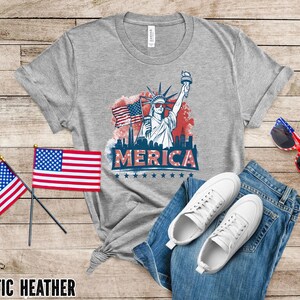 May include: Athletic heather gray t-shirt with a graphic of the Statue of Liberty, American flags, and the word "MERICA". The shirt is paired with blue jeans, white sneakers, and red, white, and blue sunglasses.