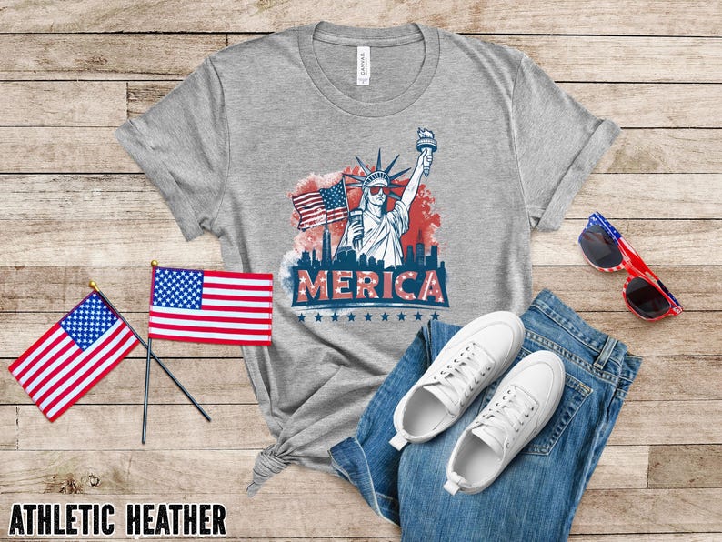 May include: Athletic heather gray t-shirt with a graphic of the Statue of Liberty, American flags, and the word "MERICA". The shirt is paired with blue jeans, white sneakers, and red, white, and blue sunglasses.