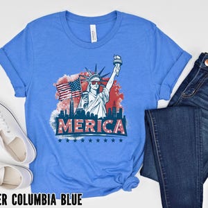 May include: A light blue t-shirt with a graphic of the Statue of Liberty holding a torch and American flag, with the word "MERICA" below. The shirt is paired with blue jeans and white sneakers. The text "HEATHER COLUMBIA BLUE" is at the bottom.