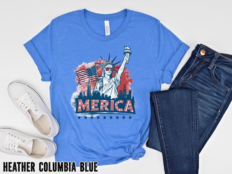 May include: A light blue t-shirt with a graphic of the Statue of Liberty holding a torch and American flag, with the word "MERICA" below. The shirt is paired with blue jeans and white sneakers. The text "HEATHER COLUMBIA BLUE" is at the bottom.