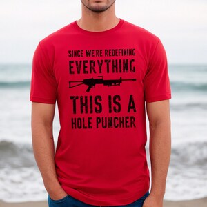 May include: Red t-shirt with a black graphic of a rifle and the text "Since we're redefining everything this is a hole puncher".