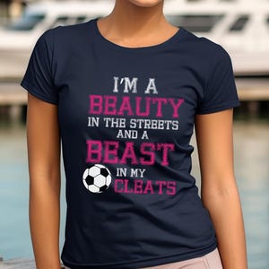 May include: Navy blue t-shirt with white text that reads "I'm a beauty in the streets and a beast in my cleats". A white soccer ball is featured below the text.