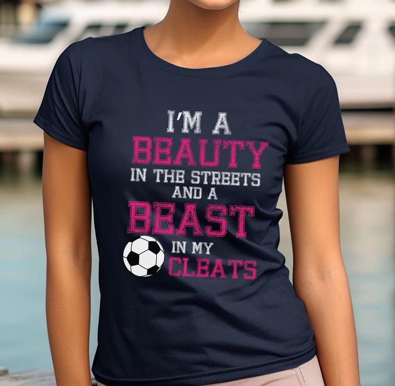 May include: Navy blue t-shirt with white text that reads "I'm a beauty in the streets and a beast in my cleats". A white soccer ball is featured below the text.