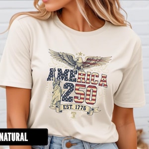 Peut inclure: T-shirt crème avec un motif patriotique. Le graphisme comprend un aigle, la Statue de la Liberté et le texte "AMERICA 250 EST. 1776" en rouge, blanc et bleu. Le t-shirt est de style col rond classique.