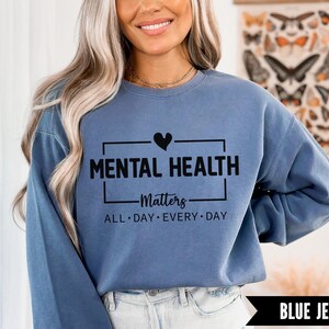 Peut inclure: Sweat-shirt bleu jean avec l'inscription noire "MENTAL HEALTH Matters ALL DAY EVERY DAY". Le motif comprend un symbole de c&oelig;ur au-dessus du mot "HEALTH".