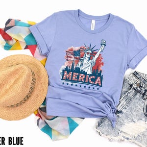 May include: Heather blue t-shirt with a graphic of the Statue of Liberty, an American flag, and the word "MERICA". The shirt is paired with denim shorts, a straw hat, and a patterned scarf.