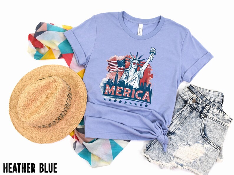 May include: Heather blue t-shirt with a graphic of the Statue of Liberty, an American flag, and the word "MERICA". The shirt is paired with denim shorts, a straw hat, and a patterned scarf.