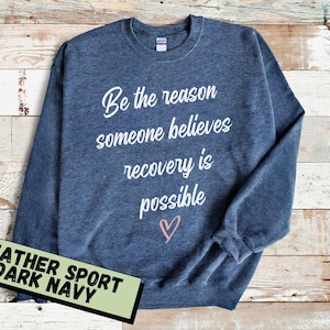 Puede incluir: Una sudadera azul marino oscuro con el texto "Be the reason someone believes recovery is possible" en escritura blanca. Un pequeño corazón rosa está debajo del texto. La sudadera está sobre un fondo de madera clara.