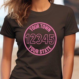 以下が含まれることがあります： 「YOUR TOWN 12345 YOUR STATE」と書かれたピンクの円形のグラフィックが施された茶色のTシャツです。