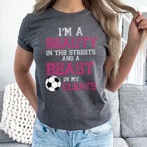 May include: A gray t-shirt with the text "I'm a beauty in the streets and a beast in my cleats" in pink lettering. A white soccer ball is centered below the text.