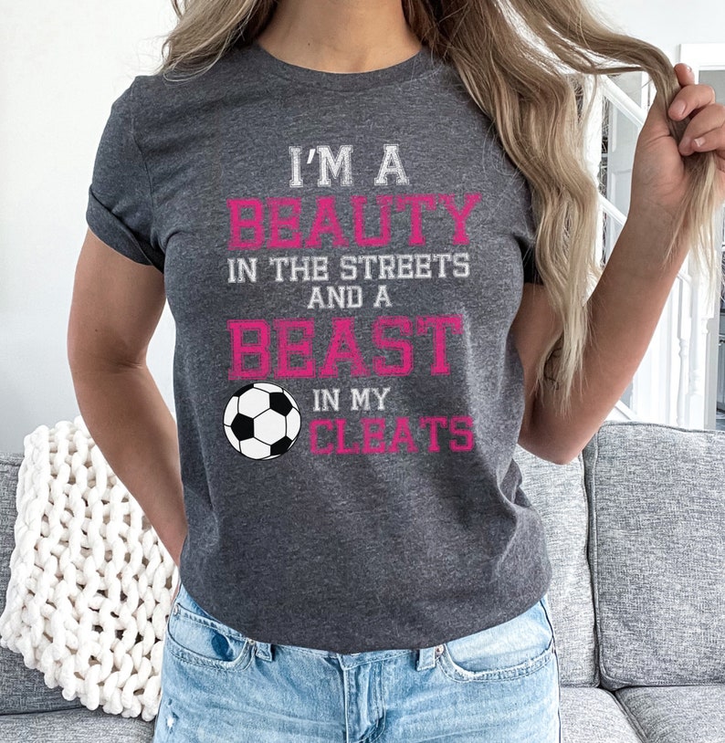 May include: A gray t-shirt with the text "I'm a beauty in the streets and a beast in my cleats" in pink lettering. A white soccer ball is centered below the text.