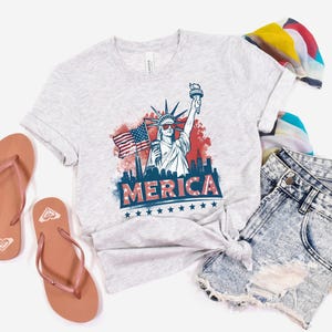 May include: Light gray t-shirt with a graphic of the Statue of Liberty holding a torch and an American flag, with the word "MERICA" in red and blue. The shirt is paired with denim shorts and brown flip-flops.