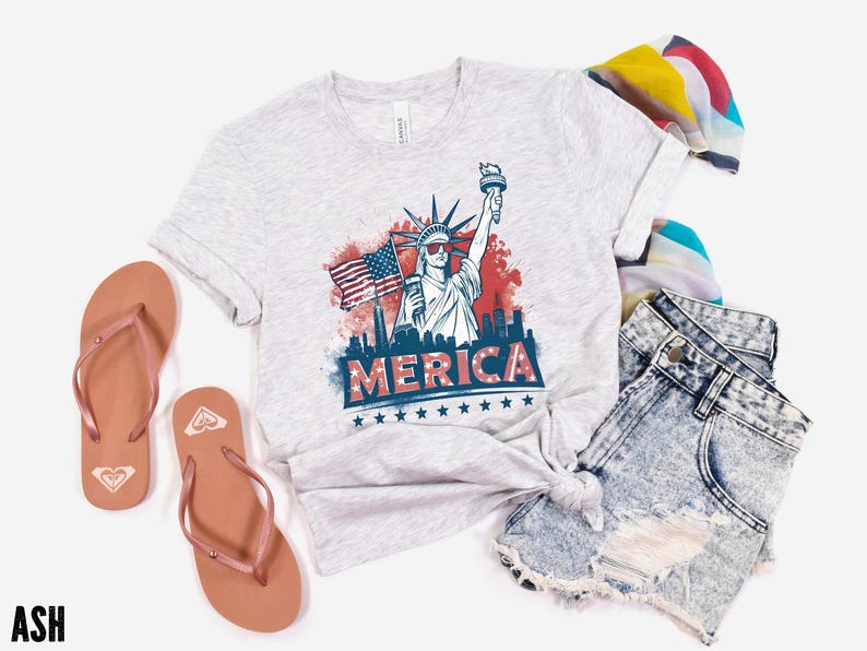 May include: Light gray t-shirt with a graphic of the Statue of Liberty holding a torch and an American flag, with the word "MERICA" in red and blue. The shirt is paired with denim shorts and brown flip-flops.
