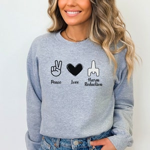 May include: A light gray sweatshirt with the words "Peace Love Harm Reduction" in black, accompanied by corresponding hand symbols and a heart. The sweatshirt is a crew neck style.