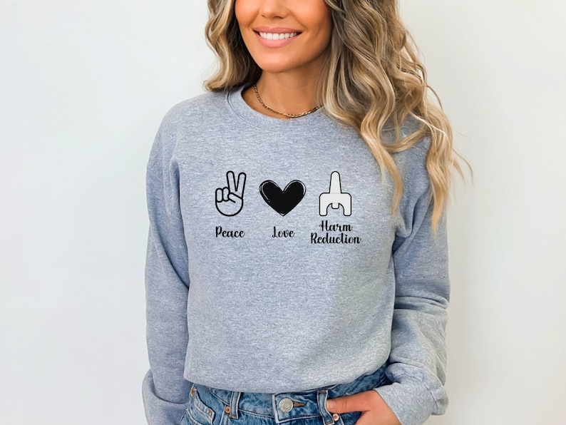 May include: A light gray sweatshirt with the words "Peace Love Harm Reduction" in black, accompanied by corresponding hand symbols and a heart. The sweatshirt is a crew neck style.