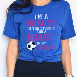 May include: A royal blue t-shirt with the text "I'm a beauty in the streets and a beast in my cleats" in white and pink lettering. A white soccer ball is centered below the text.
