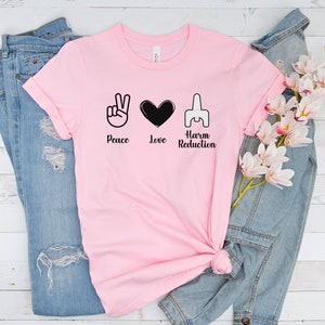 May include: A light pink t-shirt with the words "Peace, Love, Harm Reduction" and corresponding symbols in black. The shirt is displayed with a pair of blue jeans and a denim jacket, along with a sprig of white and pink flowers.