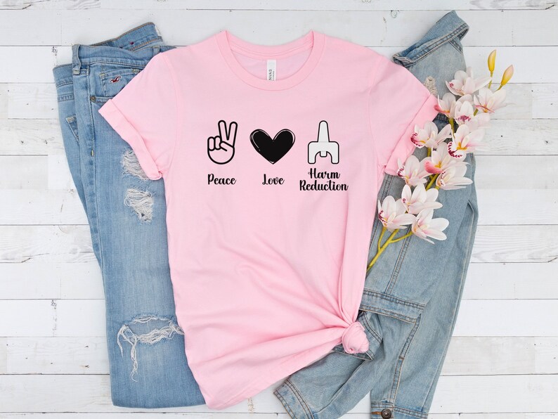May include: A light pink t-shirt with the words "Peace, Love, Harm Reduction" and corresponding symbols in black. The shirt is displayed with a pair of blue jeans and a denim jacket, along with a sprig of white and pink flowers.