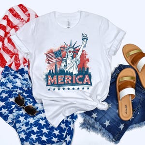 May include: White t-shirt with a graphic of the Statue of Liberty, American flag, and the word "MERICA". The shirt is paired with denim shorts, sandals, and patriotic-themed fabric.