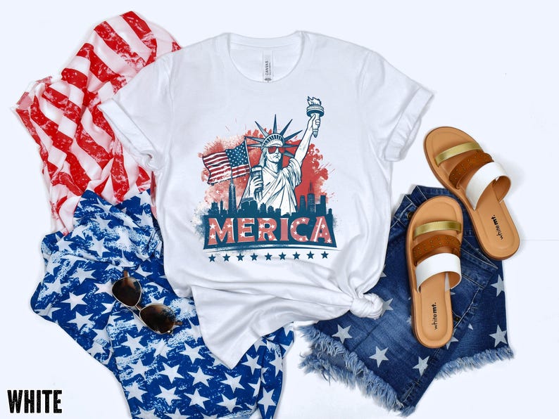 May include: White t-shirt with a graphic of the Statue of Liberty, American flag, and the word "MERICA". The shirt is paired with denim shorts, sandals, and patriotic-themed fabric.