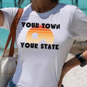 May include: White t-shirt with a retro sunset graphic and the text "YOUR TOWN YOUR STATE" printed in black.