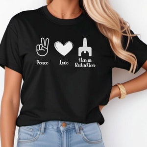May include: Black t-shirt with the words "Peace, Love, Harm Reduction" and corresponding white graphic symbols: a peace sign, a heart, and a stylized syringe. The shirt is a crew neck style.