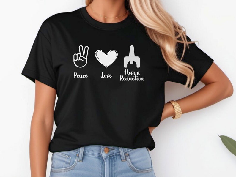 May include: Black t-shirt with the words "Peace, Love, Harm Reduction" and corresponding white graphic symbols: a peace sign, a heart, and a stylized syringe. The shirt is a crew neck style.