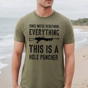 May include: A green t-shirt with a black graphic print of a rifle and the text "Since we're redefining everything, this is a hole puncher".