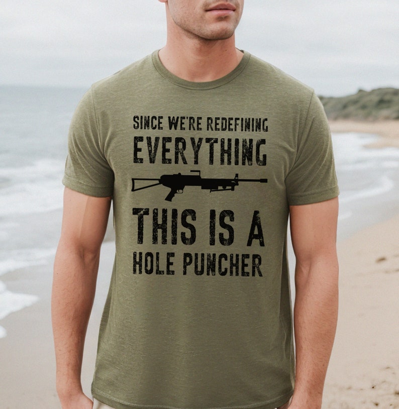 May include: A green t-shirt with a black graphic print of a rifle and the text "Since we're redefining everything, this is a hole puncher".
