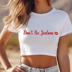 Puede incluir: Un top corto blanco con texto rojo que dice "Don't Be Jealous" con un estampado de labios rojos.