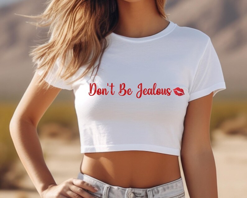 Puede incluir: Un top corto blanco con texto rojo que dice "Don't Be Jealous" con un estampado de labios rojos.