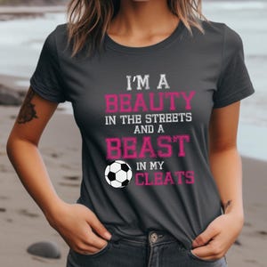 May include: A dark gray t-shirt with the text "I'm a beauty in the streets and a beast in my cleats" in pink lettering. A white soccer ball is featured below the text.