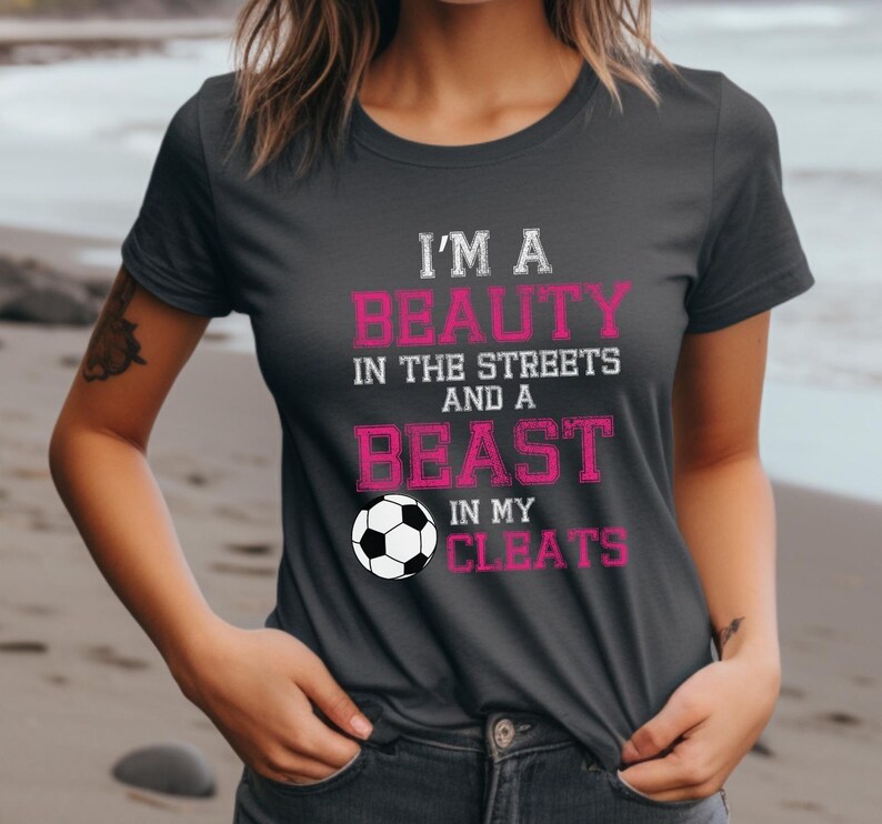 May include: A dark gray t-shirt with the text "I'm a beauty in the streets and a beast in my cleats" in pink lettering. A white soccer ball is featured below the text.