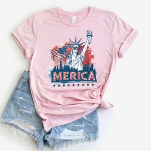 May include: Pink t-shirt with a graphic of the Statue of Liberty, an American flag, and the word "MERICA" in red, white, and blue. The shirt is paired with light blue denim shorts.