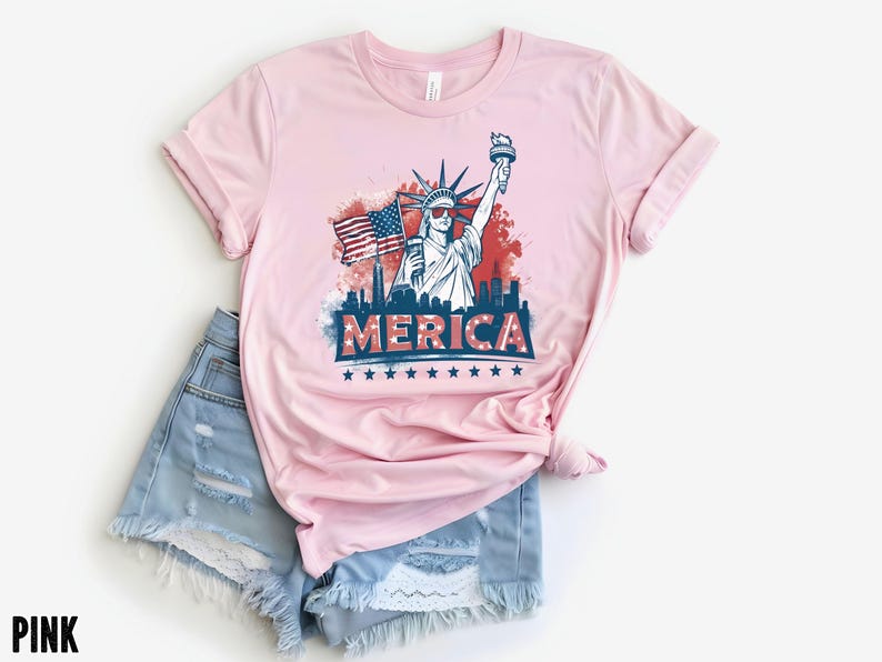 May include: Pink t-shirt with a graphic of the Statue of Liberty, an American flag, and the word "MERICA" in red, white, and blue. The shirt is paired with light blue denim shorts.