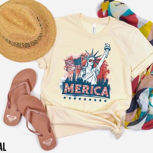 May include: Cream-colored t-shirt with a graphic of the Statue of Liberty, American flag, and the word "MERICA". The shirt is paired with a straw hat, brown sandals, and a colorful scarf. The word "NATURAL" is at the bottom.
