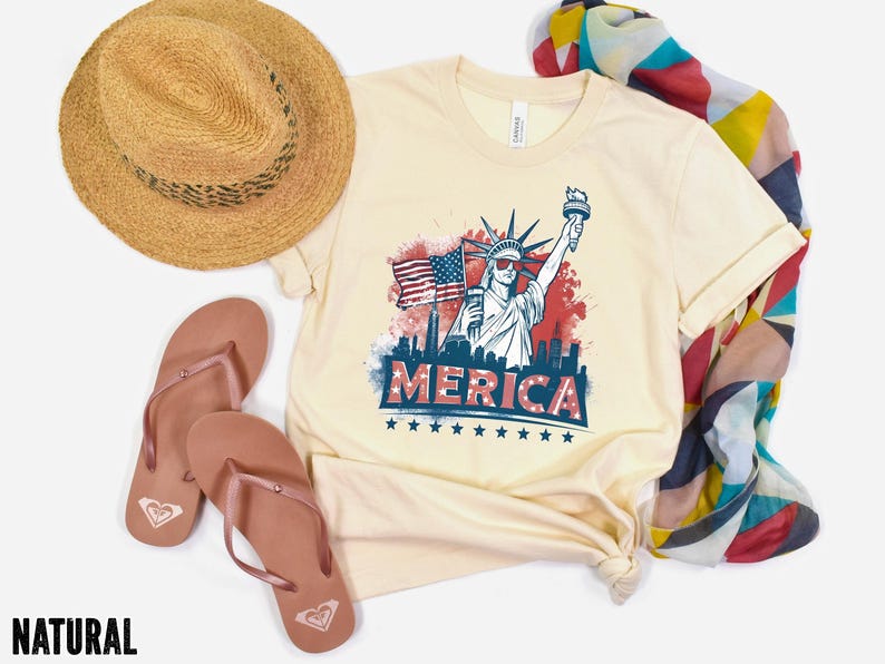 May include: Cream-colored t-shirt with a graphic of the Statue of Liberty, American flag, and the word "MERICA". The shirt is paired with a straw hat, brown sandals, and a colorful scarf. The word "NATURAL" is at the bottom.