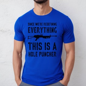 May include: A royal blue t-shirt with a black graphic print that says "Since we're redefining everything, this is a hole puncher". The graphic also includes a silhouette of a rifle.