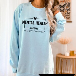 Peut inclure: Un sweat-shirt bleu clair avec le texte "MENTAL HEALTH Matters ALL DAY EVERY DAY" en noir. Un c&oelig;ur noir est au-dessus du mot "Matters". Le mot "CHAMBRAY" est dans une banni&egrave;re noire en bas.