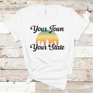 May include: White t-shirt featuring a retro sunset graphic and the text "Your Town Your State" in black.